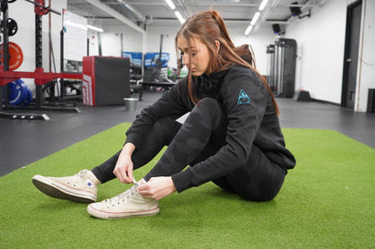 Woman sitting on the ground, tying her shoelaces. She is wearing the Collective Hoodie in Black, the logo is showing on the uppersleeve