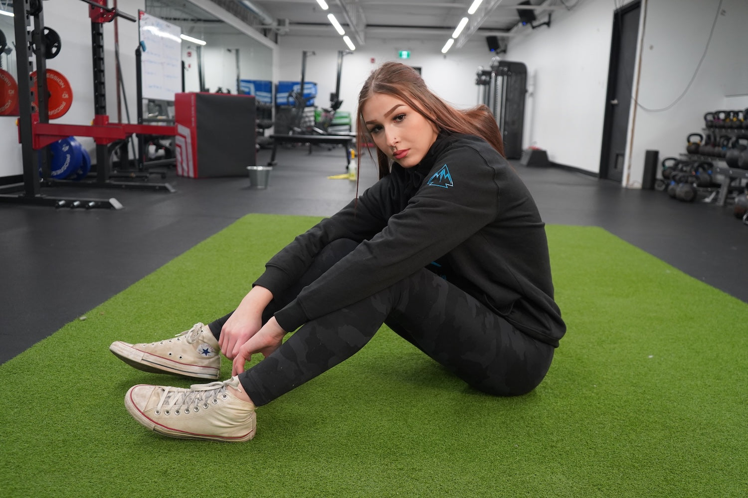 Woman sitting on the ground, looking at the camera. She is wearing the Collective Hoodie in Black, the logo is showing on the uppersleeve