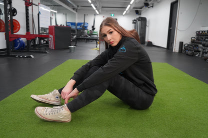 Woman sitting on the ground, looking at the camera. She is wearing the Collective Hoodie in Black, the logo is showing on the uppersleeve
