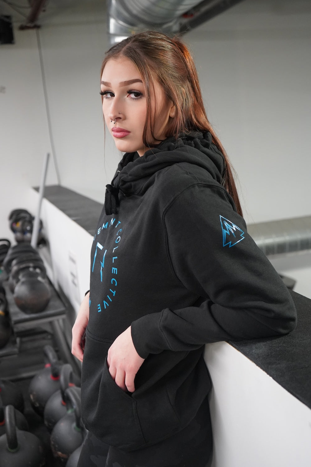 Woman standing, looking at the camera. She is wearing the Collective Hoodie in Black, the logo is showing on the uppersleeve with a little of the front decal showing