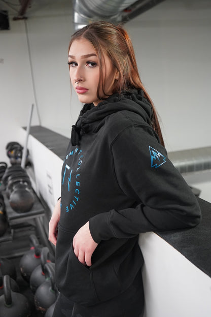 Woman standing, looking at the camera. She is wearing the Collective Hoodie in Black, the logo is showing on the uppersleeve with a little of the front decal showing