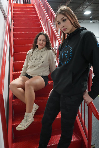 Two women in a red stairwell. One is sitting and wearing the Collective Hoodie in Tan. The other is standing and wearing the Collective Hoodie in Black