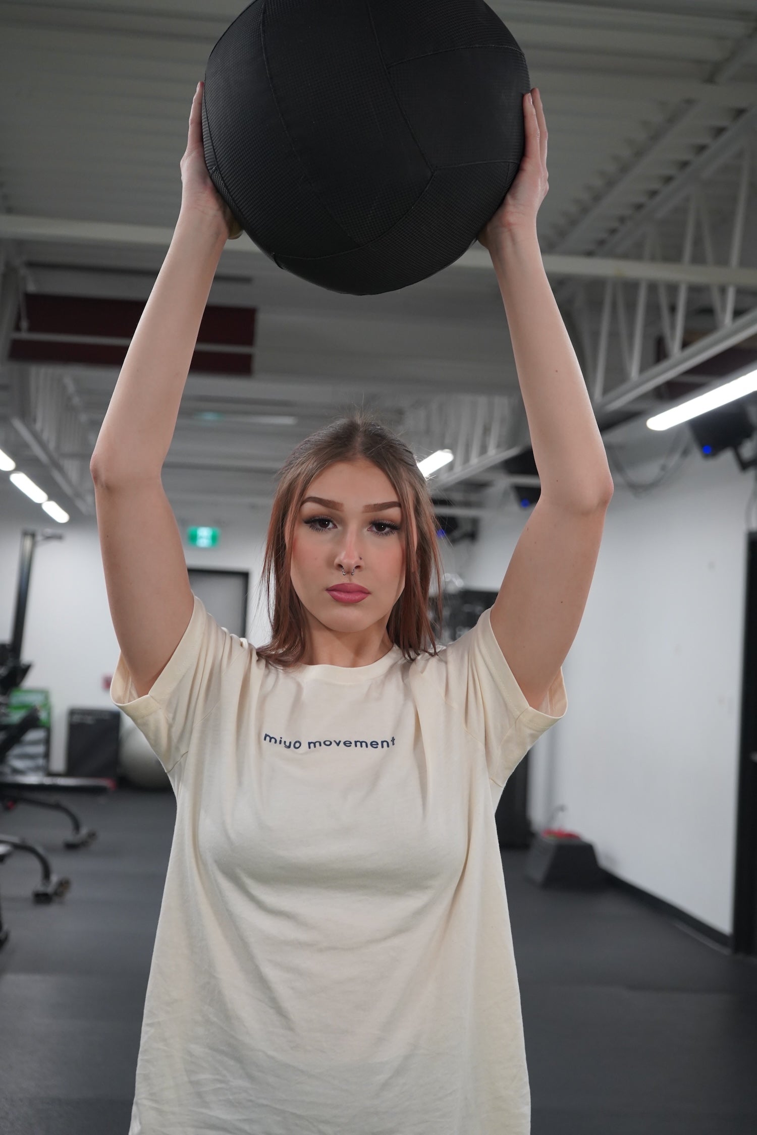 A woman holding a medicine ball above her head. She is wearing the Movement T-shirt in Natural