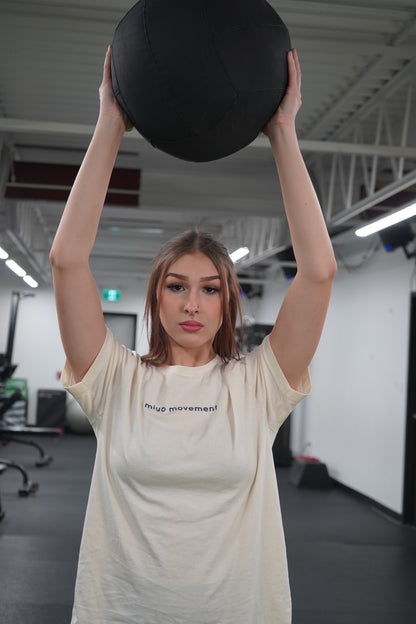 A woman holding a medicine ball above her head. She is wearing the Movement T-shirt in Natural